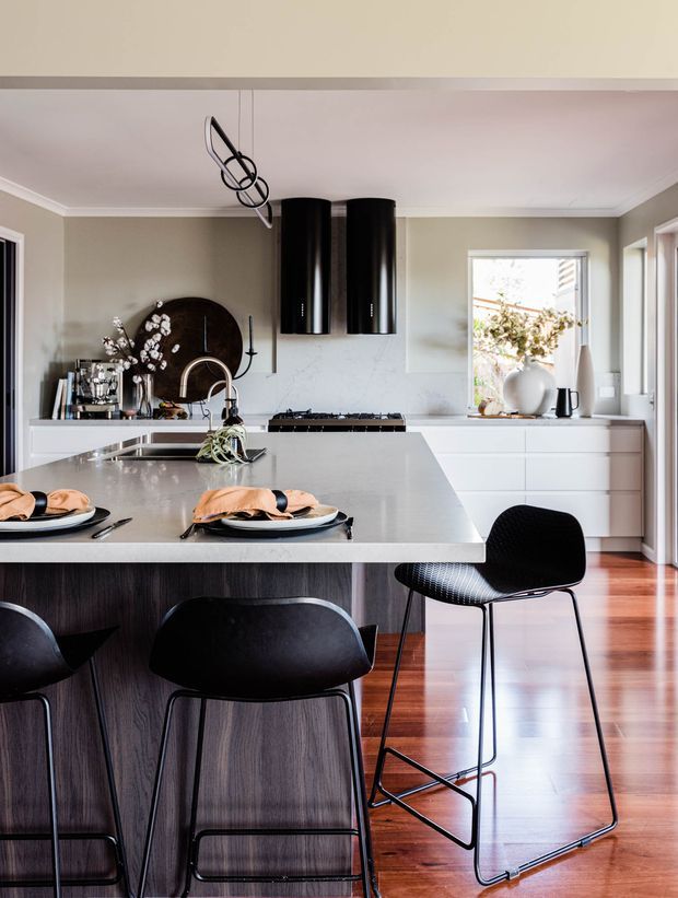 A bright, contemporary kitchen centred on a large island with a pale stone benchtop and dark timber cabinetry. The room features white cabinetry, a marble-look splashback, black cylindrical rangehoods, timber flooring, black bar stools, pendant lighting, and a window overlooking greenery.