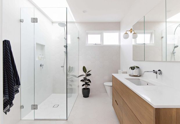 Bright contemporary bathroom with a frameless glass walk-in shower, wall-hung shower fixtures, freestanding toilet, double vanity with a pale timber cabinetry base and white benchtop, large mirrored wall cabinetry, and white hexagonal wall and floor tiles. High horizontal windows and a potted plant add natural light and greenery.