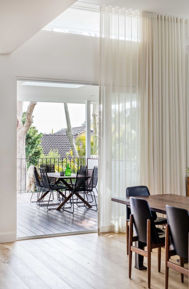 Bright, airy dining area with pale timber flooring, a dark timber dining table and black dining chairs, framed by full-height sheer curtains. Large sliding glass doors open onto a timber-decked balcony with outdoor dining furniture and leafy suburban views. A skylight and high ceiling add abundant natural light.