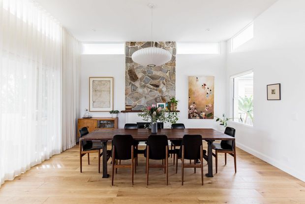 Bright, spacious dining room with a long timber dining table and dark upholstered chairs, surrounded by sheer floor-to-ceiling curtains and high horizontal windows. A prominent irregular stone feature wall, sculptural pendant light, timber sideboard, artwork, and pale timber flooring create a warm contemporary interior.
