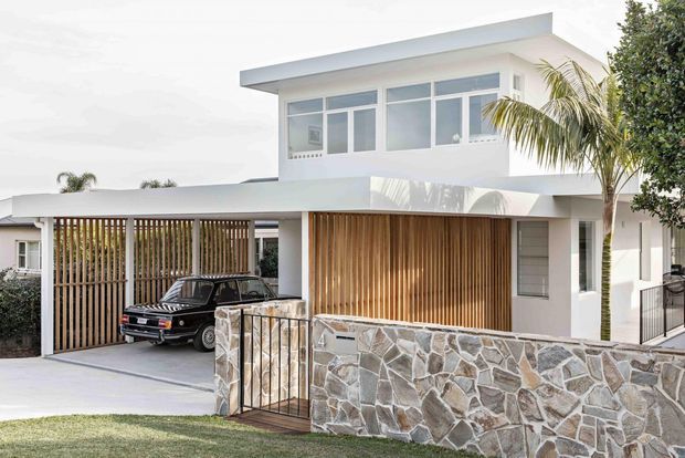 A bright contemporary two-storey home exterior with a flat roof, white rendered walls, large upper-level windows, timber vertical screens and garage/carport enclosure, a natural irregular-stone boundary wall, and a palm tree in the front landscape.