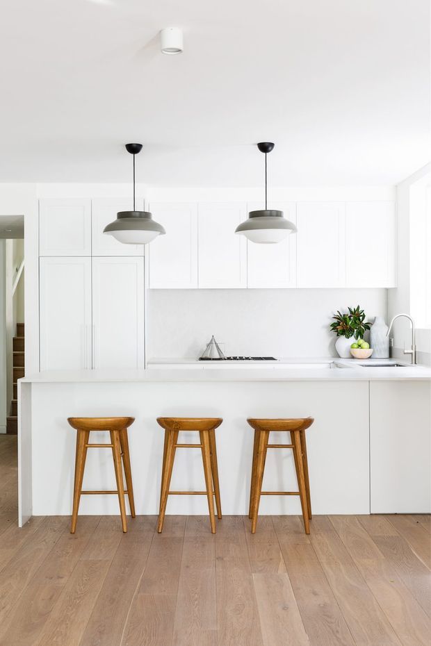 Bright, minimalist white kitchen with full-height white cabinetry, a long white island with three wooden bar stools, integrated cooktop, undermount sink and brushed metal tap. Two pendant lights hang above the island, while light timber flooring and small greenery add warmth. The room has a clean, airy contemporary feel.