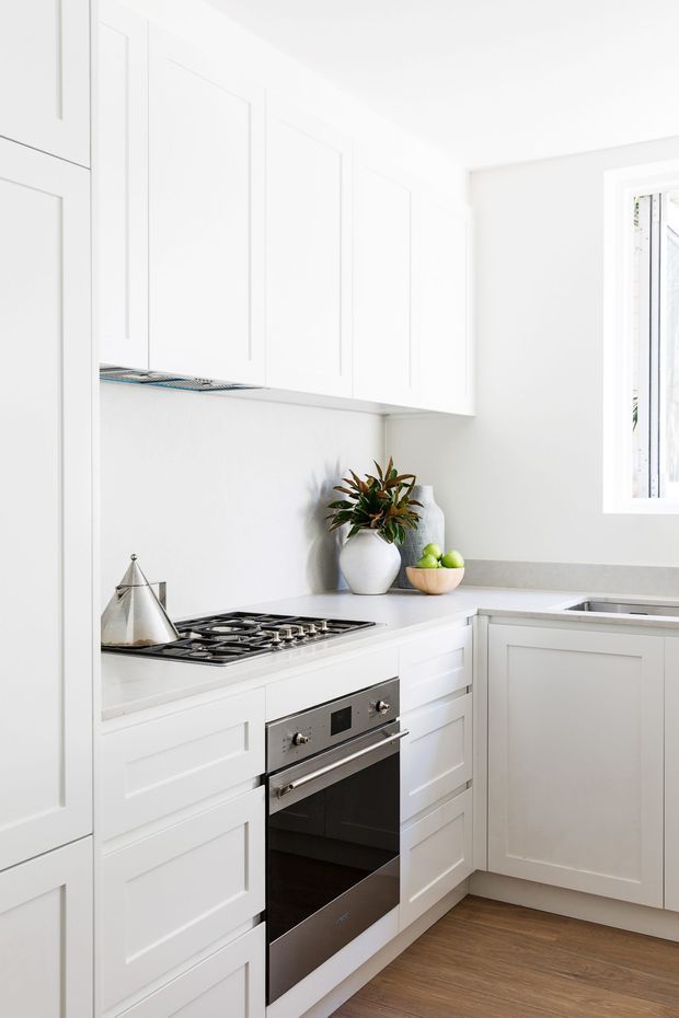 Bright, compact white kitchen with shaker-style wall and base cabinetry, a light grey benchtop, stainless-steel gas cooktop and built-in oven, integrated sink, and warm timber-look flooring. A small window provides natural light, while minimal decorative greenery and fruit add subtle warmth.