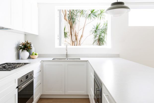 Bright, contemporary white L-shaped kitchen with shaker-style cabinetry, expansive white benchtops, integrated stainless-steel appliances, a gas cooktop, undermount sink, and a large window overlooking leafy planting. A modern pendant light hangs above the island or peninsula, with warm timber flooring providing contrast.