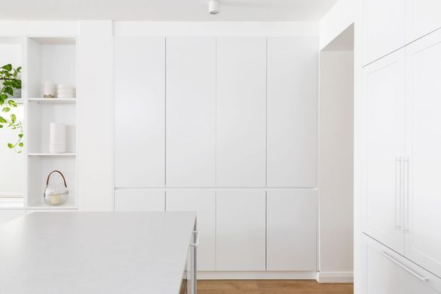 Bright, minimalist white kitchen with extensive handleless floor-to-ceiling cabinetry, integrated shelving, a pale stone island benchtop, and a timber floor. A stainless steel gooseneck tap is visible at the island, with subtle greenery and neutral styling.