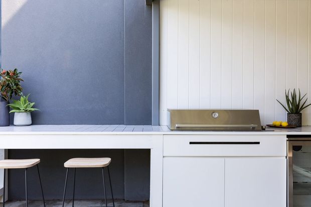 A compact contemporary outdoor kitchen or entertaining area with white vertical-panel cabinetry, a stainless-steel barbecue hood, dark grey wall cladding, a pale tiled benchtop, and two simple stools at a breakfast bar. Potted plants provide small natural accents.