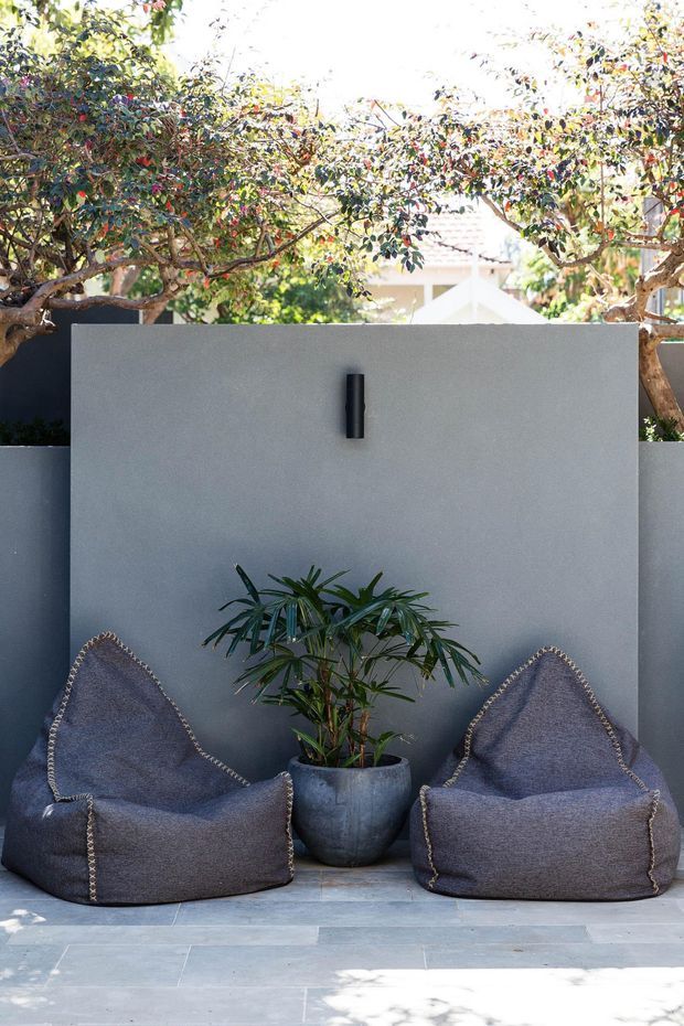 A compact outdoor seating nook with two dark grey beanbag-style lounge chairs arranged against a smooth charcoal-grey rendered wall. A potted palm sits between the chairs, with mature leafy trees overhead and pale stone paving underfoot. A simple black wall light provides a small architectural accent.