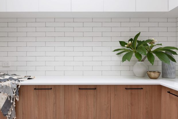 Bright contemporary kitchen with a white subway-tile backsplash, white benchtop, white overhead cabinetry, and warm timber-grain base cabinetry with black handles. A plant and decorative objects sit on the corner benchtop, while a folded textile rests at the left edge.