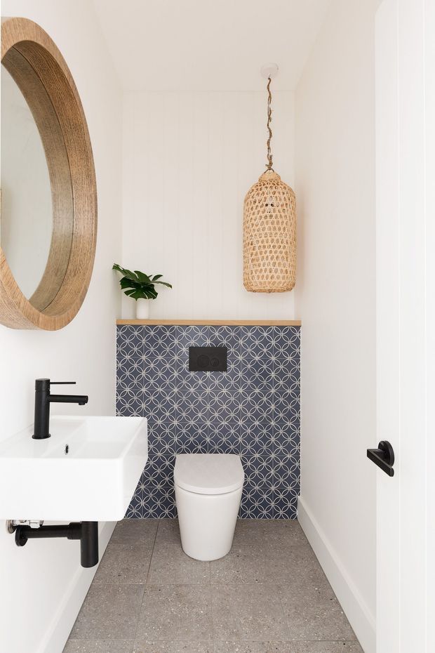 A compact contemporary bathroom with a wall-hung white toilet, white wall-mounted basin, matte black tapware and plumbing, and a patterned blue-grey tiled feature wall. Large grey terrazzo-style floor tiles, white walls, a timber-framed round mirror, a woven pendant light, and a small plant create a clean, warm, modern look.