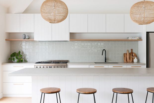 Bright contemporary kitchen with white flat-panel cabinetry, a long island with seating, pale textured tiled splashback, timber shelving, and woven pendant lights. Warm timber stools and natural accents soften the predominantly white scheme.