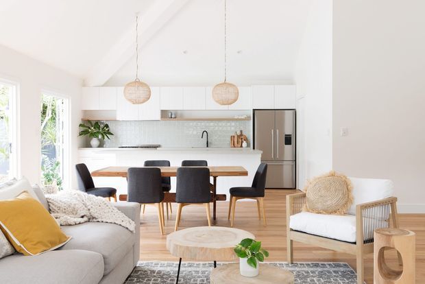 Bright open-plan kitchen, dining and living area with white cabinetry, pale timber flooring, a tiled backsplash, stainless-steel appliances and a warm timber dining table. The room is furnished with a neutral sofa, dark upholstered dining chairs, woven pendant lights, a rattan armchair and soft coastal-inspired accents. Large windows bring in abundant natural light.