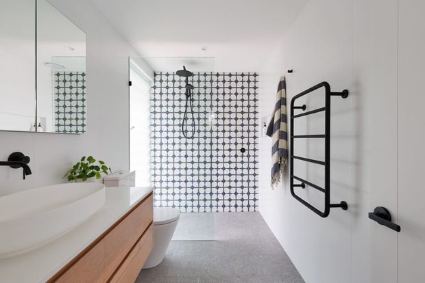 Bright contemporary bathroom with a freestanding white basin on a timber vanity, wall-mounted black tapware, a patterned tiled shower enclosure, a white toilet, and a black heated towel rail. The space has white walls, a large mirror, and a grey tiled floor.