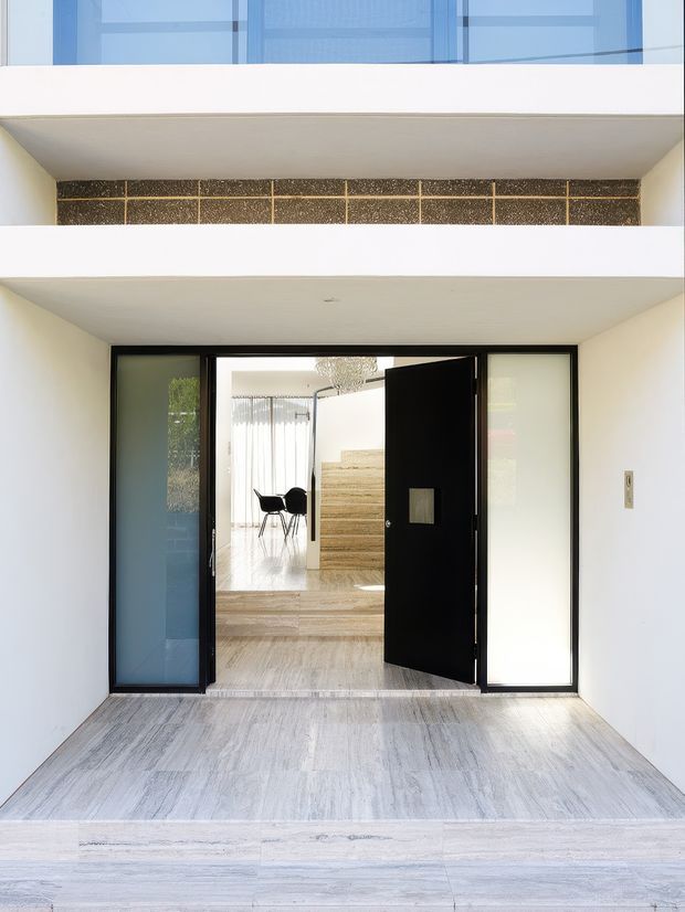 A bright, minimalist residential entrance viewed from a covered exterior vestibule. A large black pivot door with frosted and translucent sidelights opens toward a refined interior featuring pale stone flooring, a short flight of stone steps, a dining area, and a sculptural pendant light. White plaster walls and projecting horizontal balcony or canopy slabs frame the entry, with a narrow band of dark textured masonry above.