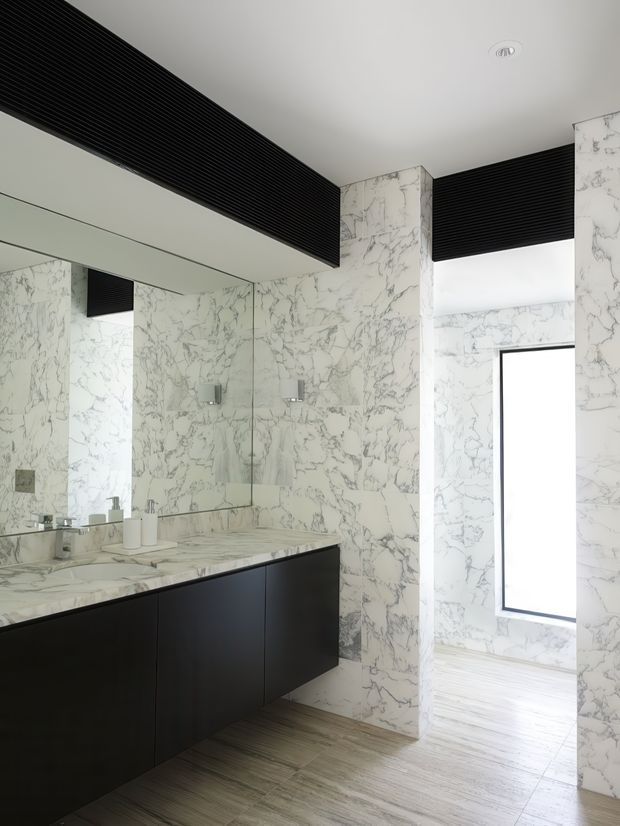 A contemporary bathroom with full-height white marble-look veined wall surfaces, a large frameless mirror, double vanity with a dark matte built-in cabinet, and a matching marble benchtop. Black linear ceiling grilles and a tall narrow window add strong contrast, while light timber-look flooring warms the space.