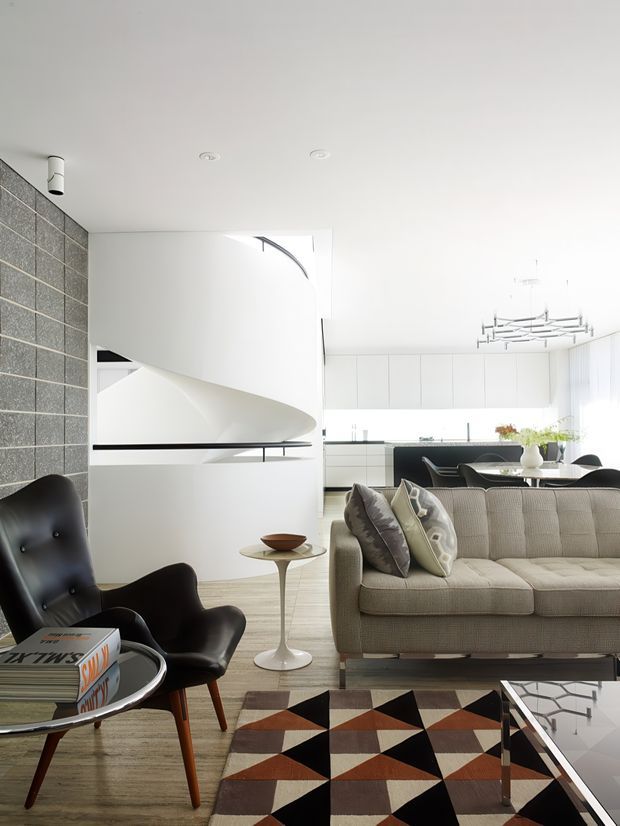 Bright, refined open-plan living area with a sculptural white curved staircase or architectural feature, a grey blockwork feature wall, contemporary kitchen cabinetry, dining area, textured neutral sofa, black leather lounge chair, geometric rug and modern pendant lighting. The space combines clean contemporary architecture with mid-century-inspired furniture and bold geometric accents.