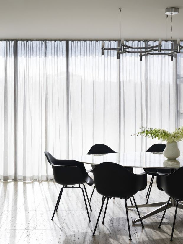 Bright, refined dining area with a large oval white dining table surrounded by black sculptural chairs. Full-height sheer white curtains create an airy backdrop, while a contemporary linear chandelier hangs overhead. The light-toned polished floor and restrained palette give the space a clean modern character.