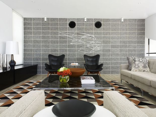 A refined contemporary living room with a dramatic grey textured block feature wall, black sculptural lounge chairs, a beige sofa, low black entertainment cabinetry, and a glass coffee table. A bold geometric patterned rug adds black, white, brown, and terracotta tones, while recessed ceiling lights and minimalist sculptural wall art complete the space.