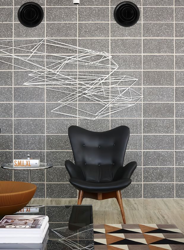 A stylish mid-century-inspired sitting area with a black sculptural lounge chair centered against a dark grey speckled grid-tile feature wall. The wall includes two black circular vents and a white geometric wire artwork. A small glass side table, sculptural wooden bowl, stacked magazines, reflective coffee table, and geometric patterned rug complete the moody, graphic interior.