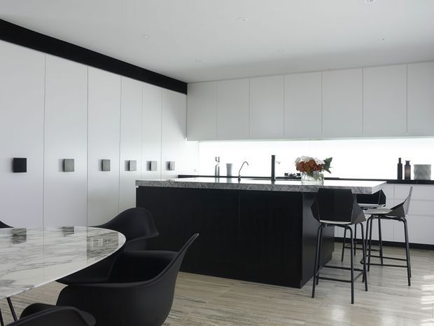 A sleek contemporary open-plan kitchen and dining area with extensive white flat-panel cabinetry, a black island and benchtop, integrated appliances, and a marble-topped dining table. Black dining chairs and bar stools contrast with pale timber-look flooring, while under-cabinet lighting creates a clean, refined atmosphere.
