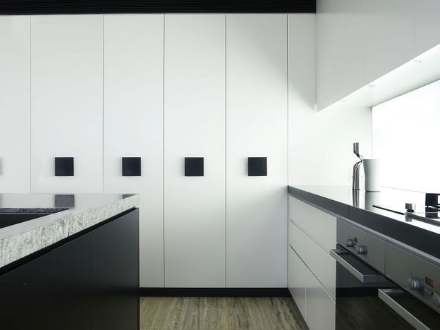 Minimalist contemporary kitchen with full-height white cabinetry, matte black square handles, dark benchtops, integrated appliances, and a central island with a pale textured stone surface. The space has strong black-and-white contrast and streamlined built-in joinery.