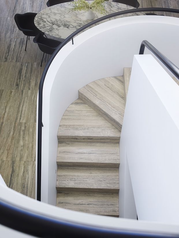 A modern curved staircase viewed from above, with pale stone or stone-look treads, smooth white curved walls, and a dark rounded handrail. A dining area with a round veined stone table and dark chairs is visible beyond the stair opening. The composition is clean and contemporary with strong sculptural curves and neutral finishes.