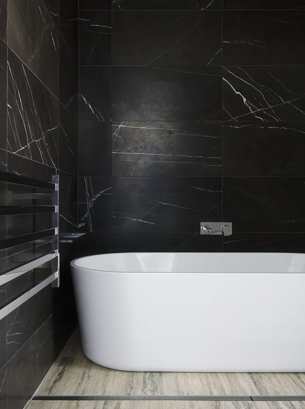 A contemporary bathroom featuring a freestanding white bathtub against dramatic black marble-effect wall tiles with white veining. A chrome wall-mounted tap is positioned above the tub, with a horizontal heated towel rail on the left. The floor has a light stone or timber-look finish, creating contrast with the dark walls.
