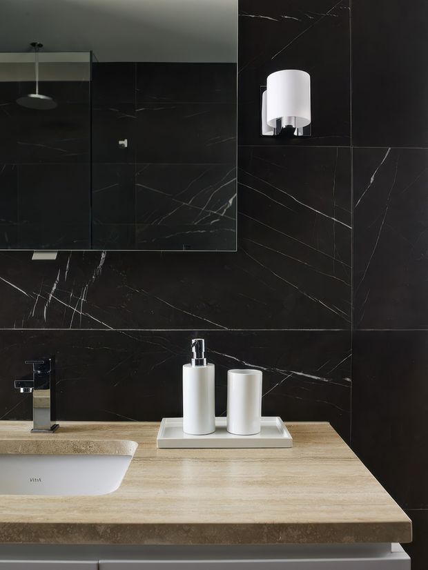 Close-up view of a refined bathroom vanity. Large dark black stone-look wall tiles with prominent white veining form the backsplash and surrounding walls. A light beige stone or engineered-stone countertop supports a white inset basin, chrome tapware, and a white tray with matching cylindrical bathroom accessories. A large mirror and wall sconce are visible above the vanity.