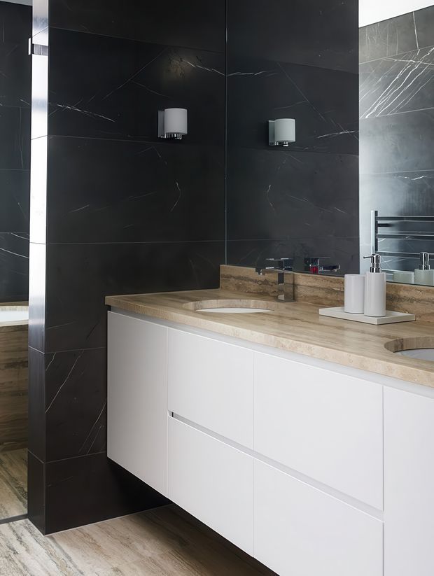 Contemporary bathroom vanity with a long white floating cabinet, double integrated basins, a beige stone benchtop, large dark marble-look wall tiles, wall sconces, and a mirrored side wall. The space has a refined, high-contrast monochromatic palette with warm stone and timber-look flooring.