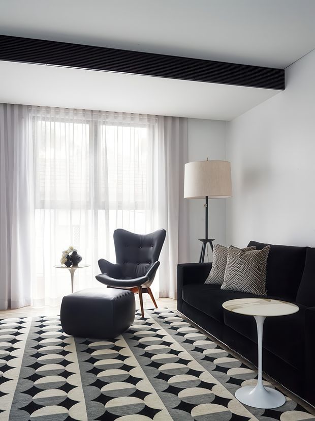 A refined living room with a black sofa, sculptural lounge chair and ottoman, geometric black-and-cream rug, slim white side table, and a tripod floor lamp. Sheer curtains provide soft daylight, while a long black linear ceiling vent adds a contemporary architectural detail.