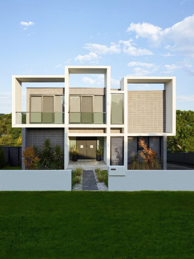 Front exterior view of a contemporary multi-storey residence with strong geometric white rectangular volumes, recessed balconies, textured masonry screens, dark window and door elements, and landscaped entry planting. A low white boundary wall and lawn frame the modern facade beneath a bright blue sky.