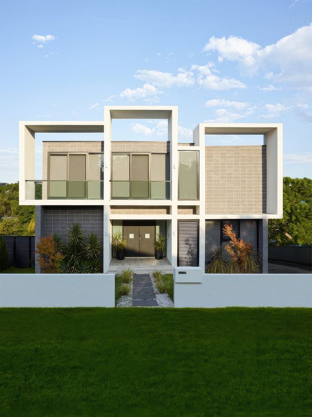 Front exterior view of a contemporary multi-level residential building with strong geometric box forms, white rendered walls, recessed balconies, patterned screening, dark window and door elements, and landscaped planting behind a low boundary wall. The facade is photographed in bright daylight against a blue sky.