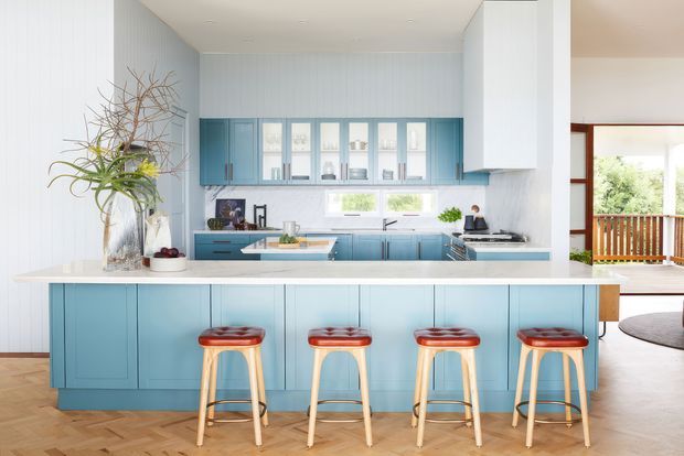 Bright, airy kitchen with a long island and four timber stools. The room features blue Shaker-style cabinetry, white benchtops, pale vertical wall panelling, timber flooring, and a bank of windows above the rear bench. A glazed opening connects to a covered outdoor deck or balcony.