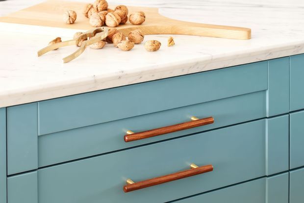 Close-up of a blue painted kitchen island or cabinet run with flat-panel drawers and warm wood-and-brass handles. A light marble-look benchtop sits above the cabinetry, with a timber chopping board and walnuts arranged on top.
