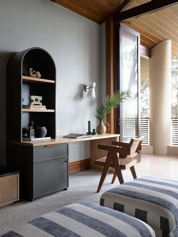 A stylish home office or study nook with a built-in dark arched shelving unit and filing drawers, a timber desk surface, sculptural wooden chair, and a small desk lamp. The room features pale textured walls, warm timber ceiling and trim, carpeted flooring, and expansive glazing opening to a covered balcony or terrace. Upholstered striped seating occupies the foreground.