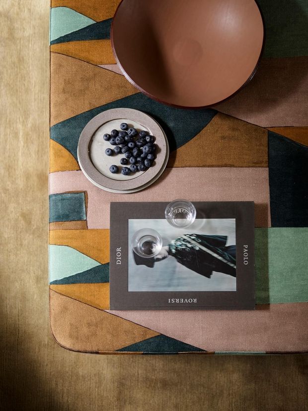 Top-down still life of a styled tabletop or dining surface. A richly patterned geometric textile or rug in ochre, muted teal, dusty pink, and tan covers most of the surface, with a large terracotta-toned bowl, a small plate of blueberries, a clear drinking glass, and a fashion book arranged on top. The scene has warm, earthy, editorial styling.