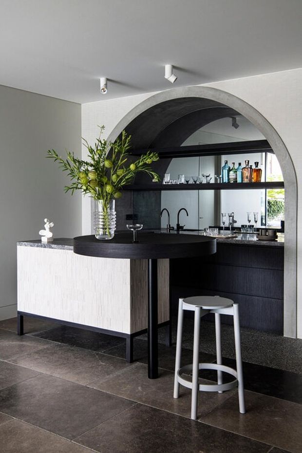 Contemporary bar and kitchen area featuring a black rounded island with integrated seating, dark cabinetry, open shelving stocked with bottles and glassware, and a large arched opening framed in textured light-grey plaster. A contrasting white textured island face, dark tiled flooring, and sculptural greenery complete the monochromatic interior.