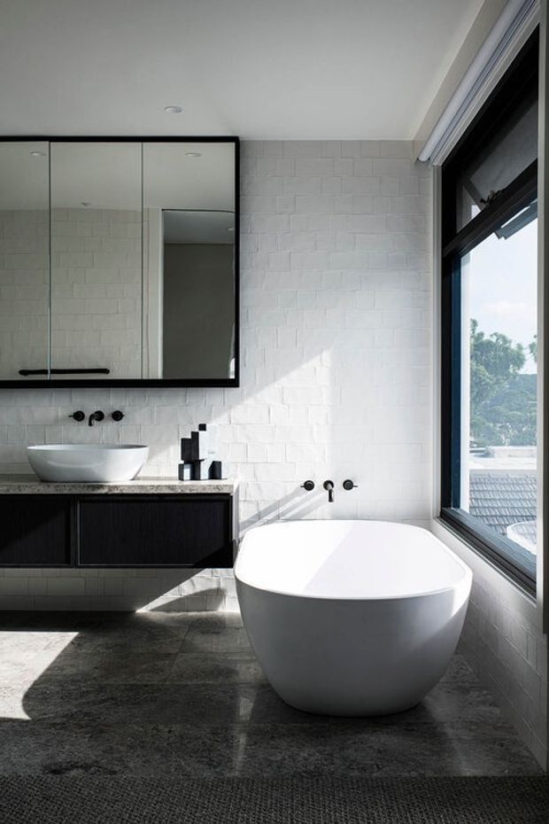 A bright contemporary bathroom featuring a freestanding white bathtub beside a large black-framed window, a floating dark vanity with a white vessel basin, mirrored wall cabinetry, white textured brick-style wall tiles, dark stone-look floor tiles, and matte black tapware. Strong natural light creates crisp shadows across the room.