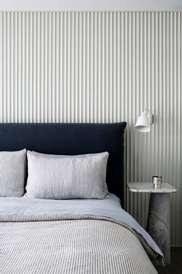 A close-up view of a refined bedroom with a dark navy upholstered headboard, light grey and white striped bedding and pillows, and a sculptural grey stone bedside table. The wall behind the bed features full-height vertical fluted or ribbed panelling, complemented by a white wall-mounted reading lamp. The palette is cool, monochromatic, and softly textured.