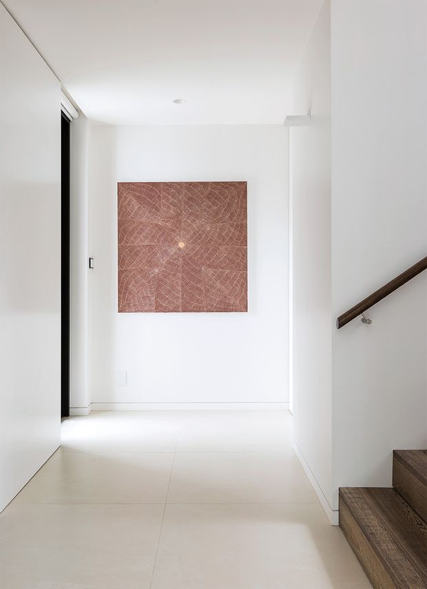 Bright minimalist interior hallway and stair landing with white walls, a pale tiled floor, a dark timber stair handrail and timber stair treads. A large square terracotta-toned abstract artwork provides the main visual accent at the end of the corridor.