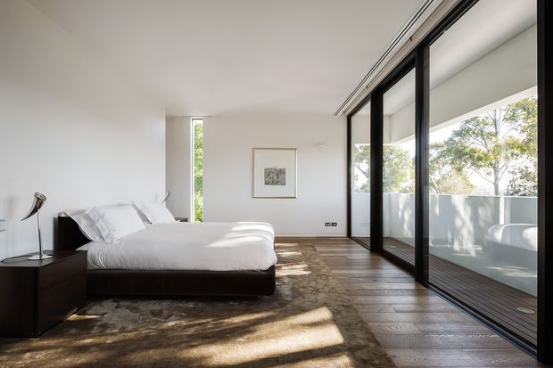 A minimalist contemporary bedroom with a low-profile dark bed, crisp white bedding, white walls, and warm dark timber flooring. Full-height black-framed sliding glass doors open onto a balcony or terrace, bringing in abundant natural light and views of trees. A large neutral rug, simple artwork, and restrained furnishings create a calm, refined atmosphere.