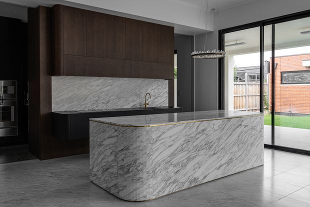 Contemporary kitchen featuring a large sculptural island clad in heavily veined white marble, dark timber cabinetry, and a matching marble splashback. A slim linear pendant hangs above the island, while full-height black-framed glazing opens to a small rear courtyard and brings in natural light.