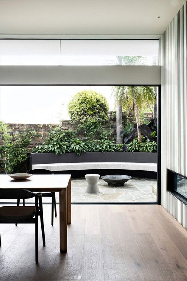 A minimalist dining area opens through full-height glazing to a landscaped courtyard. The interior features pale timber flooring, a simple dark-edged dining table with black chairs, a white sculptural stool, and a low dark bowl. The courtyard has a curved built-in bench, stone paving, brick boundary walls, dense planting, and palm trees. A vertical slatted wall and concealed roller blinds reinforce the clean contemporary design.