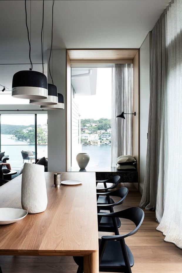 Contemporary dining area with a long light timber dining table, black dining chairs, sculptural black-and-white pendant lights, and full-height sheer curtains. A large window and glazed opening provide views across a calm waterfront setting, while warm timber detailing frames the window and a dark stone ledge sits beneath it.