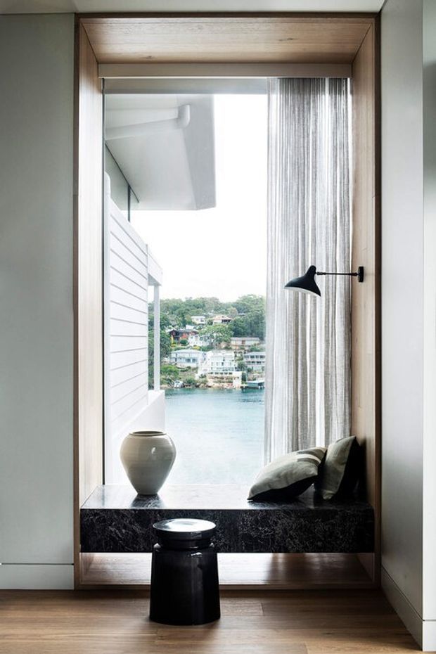 A refined interior window seat overlooking a calm waterfront scene. The deep-set window is framed in warm timber, with sheer curtains, a dark stone or marble sill/bench, neutral cushions, a black wall-mounted reading lamp, and a small black side table on a timber floor. The composition is minimalist and highly tailored, with strong natural light and a view across the water to residential buildings.