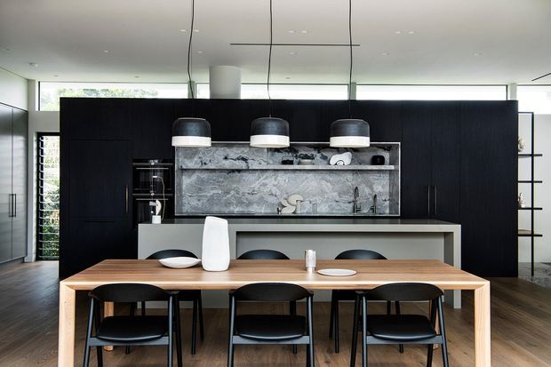 Contemporary open-plan kitchen and dining area with a long light timber dining table and black chairs in the foreground. Behind it is a dark full-height kitchen wall with integrated appliances, a pale stone island or counter, a dramatic grey stone splashback, and three large pendant lights. The room has dark timber flooring, minimal detailing, and expansive glazing that brings in natural light.