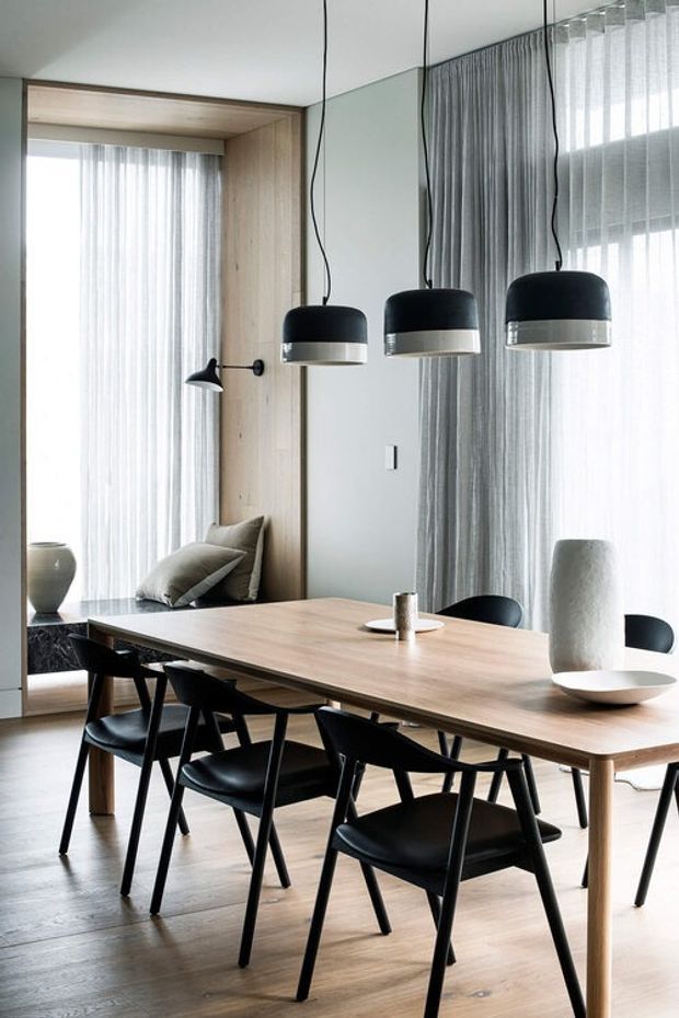 A refined contemporary dining room with a long light timber dining table surrounded by black chairs. Three black pendant lights hang above the table, while full-height sheer curtains, a large wall mirror, pale timber wall lining, and soft grey finishes create a bright, airy interior. Minimal ceramic objects and a built-in window seat add understated styling.