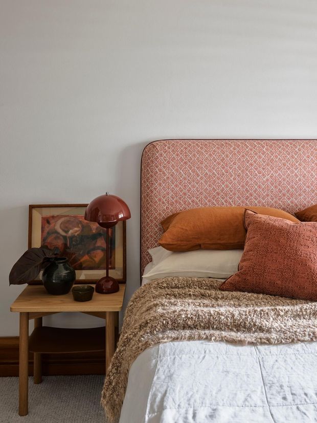 A warmly styled bedroom vignette featuring a bed with a patterned terracotta and blush upholstered headboard, rust-toned pillows, a textured brown throw, and white bedding. A timber bedside table holds a dark ceramic vase, small bowl, and burgundy dome lamp, with framed artwork leaning against the softly painted wall.