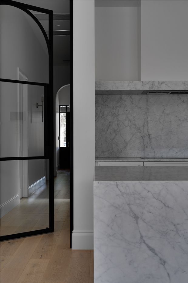 A refined contemporary kitchen viewed from an adjacent hallway, featuring a large white marble island and matching marble splashback, pale cabinetry, and a dark integrated cooktop. A black steel-framed glazed door with an arched upper detail separates the kitchen from the timber-floored corridor. The palette is monochromatic and sophisticated, with strong veining in the marble and dark metal accents.