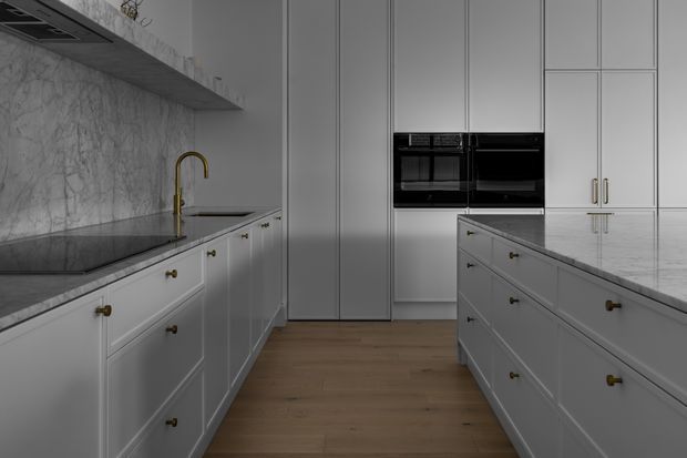 A refined contemporary kitchen with white Shaker-style cabinetry, a large central island, pale marble or stone benchtops and splashback, integrated black appliances, timber flooring, and a brass gooseneck tap. The layout is spacious and symmetrical, with tall floor-to-ceiling cabinetry and understated decorative hardware.