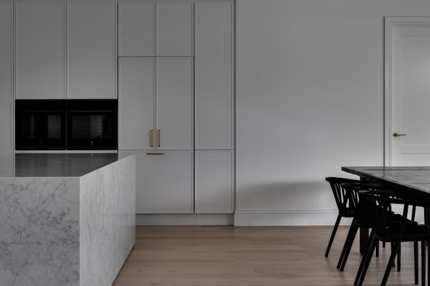 A refined contemporary kitchen and dining area with tall light-grey panelled cabinetry, integrated black appliances, a large pale marble island with waterfall edge, warm timber flooring, and a black dining table with chairs. A white panelled interior door and restrained detailing contribute to the minimalist aesthetic.
